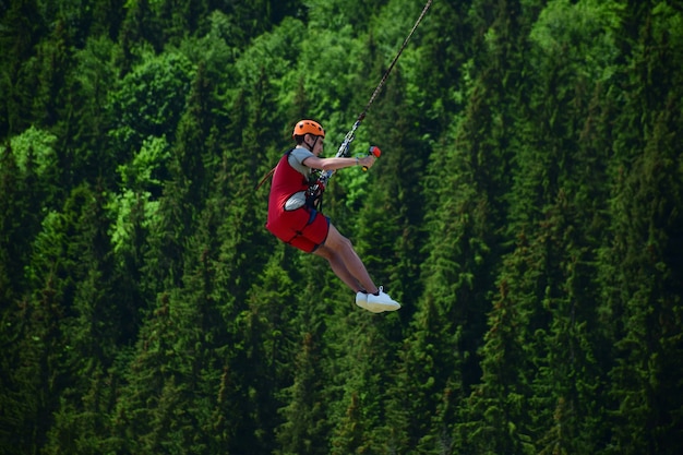 Un jeune homme portant un casque a sauté du saut à l'élastique et est maintenant suspendu à une corde, se balançant et se filmant sur une caméra vidéo de sport sur fond flou d'une forêt verte