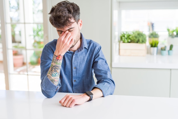 Jeune homme portant une chemise décontractée assis sur une table blanche fatigué se frottant le nez et les yeux ressentant de la fatigue et des maux de tête Concept de stress et de frustration