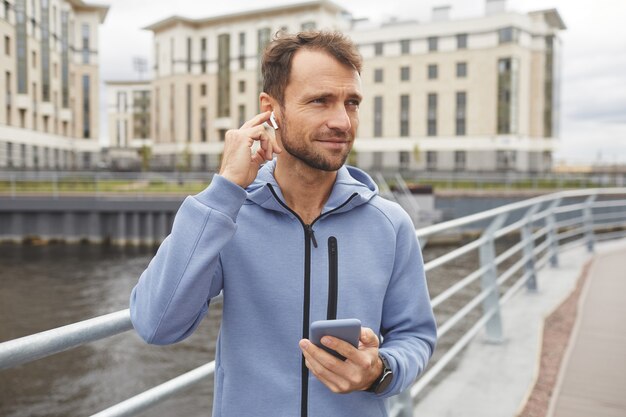Jeune homme portant des écouteurs sans fil à l'aide de son téléphone portable pour écouter de la musique lors de sa promenade dans la ville