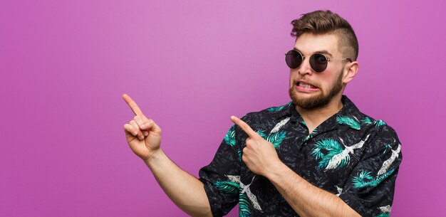 Jeune homme portant un look de vacances choqué pointant avec des index vers un espace de copie.