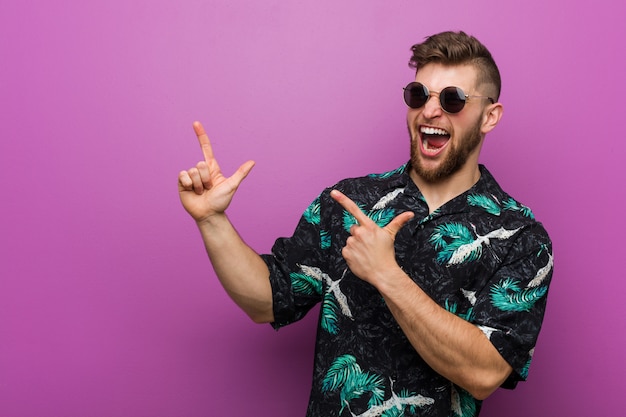 Jeune homme portant un look de vacances pointant avec des index vers un espace de copie, exprimant l'excitation et le désir.