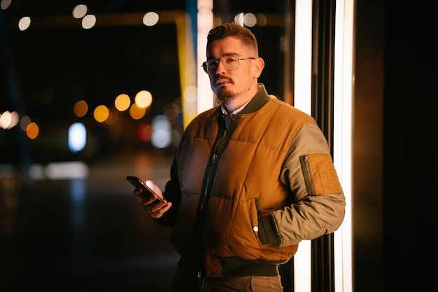 Jeune homme portant des lunettes la nuit regardant droit devant tenant son téléphone dans sa main en hiver