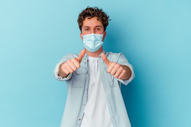 Jeune homme portant un masque antiviral isolé sur un mur bleu avec des pouces vers le haut, des acclamations pour quelque chose, le soutien et le respect du concept