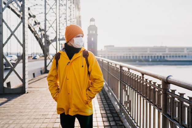 Jeune homme portant un masque chirurgical médical, un bonnet orange et un coupe-vent jaune à la recherche d'inquiétude, pensant au coronavirus pandémique, debout sur le pont dans une ville vide. Concept de virus Covid-19.