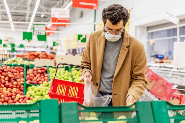 Un jeune homme portant un masque médical choisit des fruits dans un grand supermarché. Précautions lors de la pandémie de coronavirus. Alimentation équilibrée.