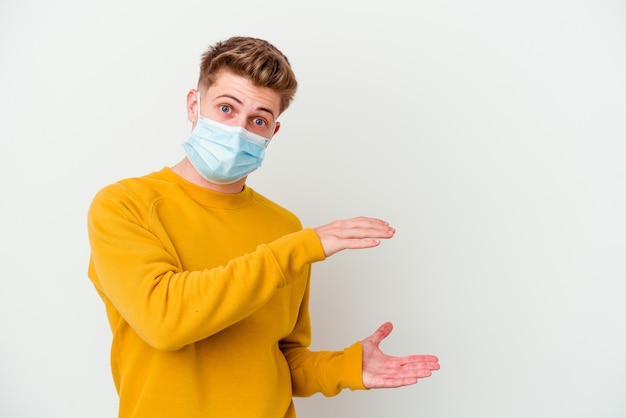 Jeune homme portant un masque pour le coronavirus isolé sur un mur blanc choqué et étonné tenant un espace de copie entre les mains.