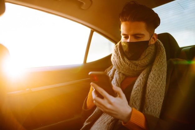 Jeune homme portant un masque stérile de protection dans une voiture de taxi sur un siège arrière vérifiant son téléphone portable.