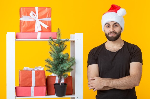 Jeune homme positif avec une barbe et un chapeau de père Noël est debout près de stand avec des cadeaux et Noël