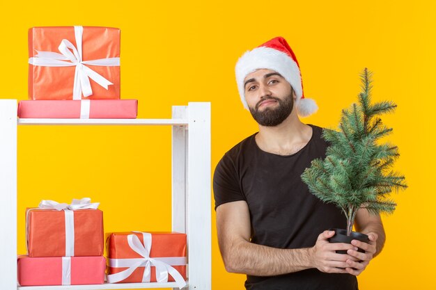 Jeune homme positif avec une barbe et un chapeau de père noël se tient près du stand avec des cadeaux et noël