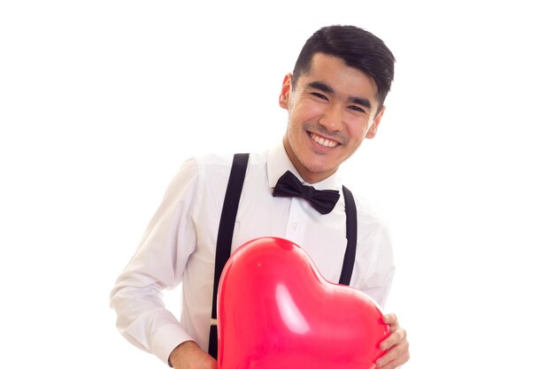 Jeune homme positif en chemise blanche avec noeud papillon noir et bretelles tenant un ballon en forme de coeur rouge