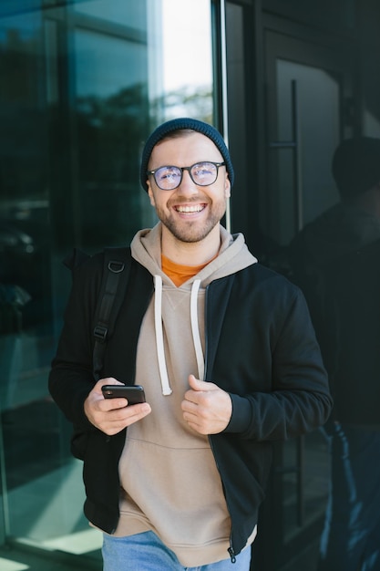 Jeune homme positif tenant un smartphone dans ses mains en regardant la caméra debout dans la rue Homme barbu à lunettes porte des vêtements décontractés Concept d'utilisation du téléphone portable