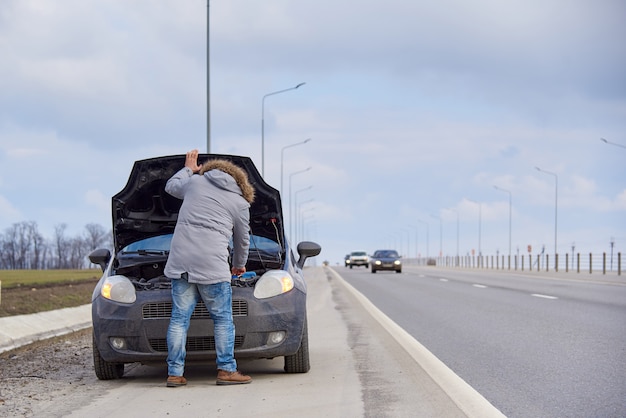 Un jeune homme près de la voiture avec un capot ouvert sur le bord de la route.