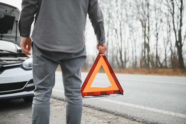 Un jeune homme près d'une voiture cassée avec un capot ouvert sur le bord de la route