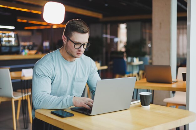 Un jeune homme prometteur en vêtements décontractés est assis à une table et travaille sur un ordinateur portable Espace de travail pour le travail à distance