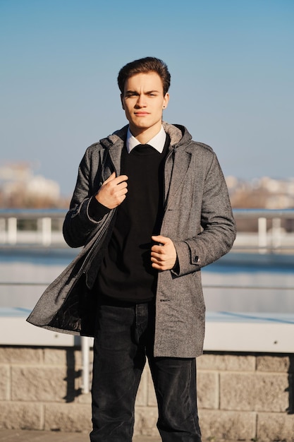 Jeune homme en pull noir, chemise blanche et jean noir tient le sol du manteau dans la rue