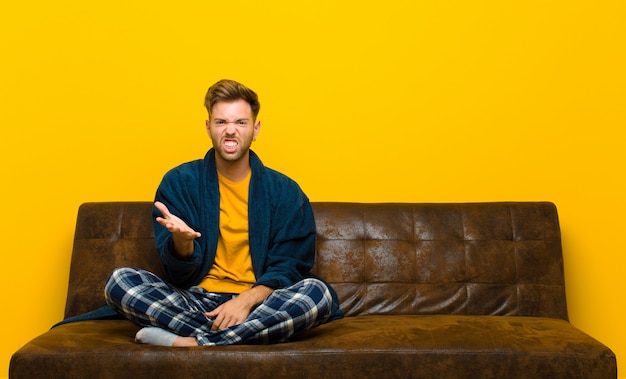 Jeune homme en pyjama qui a l'air fâché, agacé et frustré, hurlant wtf ou ce qui ne va pas avec vous. assis sur un canapé