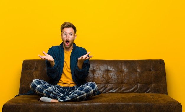 Jeune homme en pyjama se sentant extrêmement choqué et surpris, anxieux et paniqué, avec un regard stressé et horrifié. assis sur un canapé