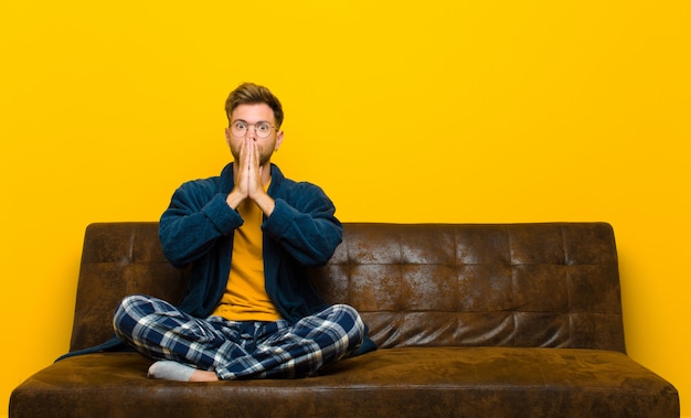 Un jeune homme en pyjama se sentant inquiet, bouleversé et effrayé, il se couvre la bouche avec les mains, a l'air inquiet et s'est perdu. assis sur un canapé