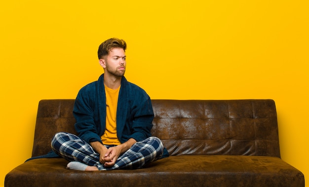 Jeune homme en pyjama se sentant triste, bouleversé ou en colère et regardant de côté avec une attitude négative, les sourcils froncés par le désaccord. assis sur un canapé