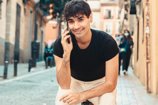 Un jeune homme de race blanche adulte assis à l'extérieur sur un pivot souriant tout en faisant un appel téléphonique et en regardant la caméra. De vraies personnes copient l'espace sur toute la longueur. Espace de copie, arrière-plan flou