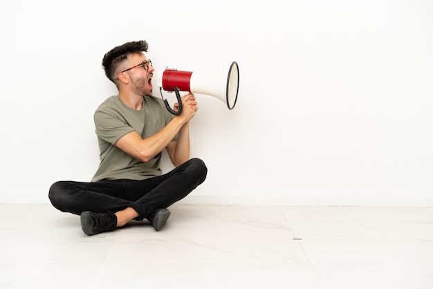 Jeune homme de race blanche assis sur le sol isolé sur fond blanc criant à travers un mégaphone