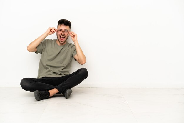 Jeune homme de race blanche assis sur le sol isolé sur fond blanc avec des lunettes et surpris