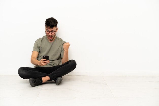 Jeune homme de race blanche assis sur le sol isolé sur fond blanc surpris et envoyant un message