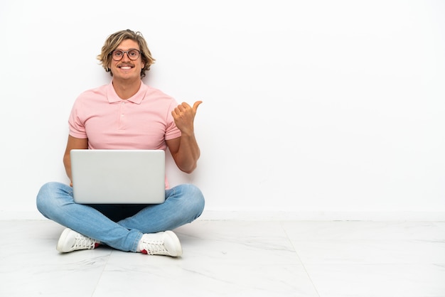 Jeune homme de race blanche assis sur le sol avec son ordinateur portable isolé sur un mur blanc pointant vers le côté pour présenter un produit