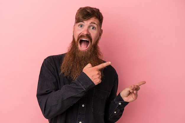 Jeune homme de race blanche au gingembre avec une longue barbe isolé sur fond rose excité pointant avec les index loin.