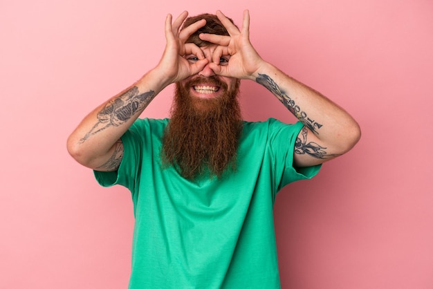 Jeune homme de race blanche au gingembre avec une longue barbe isolé sur fond rose montrant un signe d'accord sur les yeux