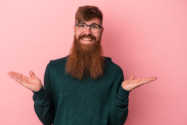 Un jeune homme de race blanche au gingembre avec une longue barbe isolée sur fond rose fait de l'échelle avec les bras, se sent heureux et confiant.