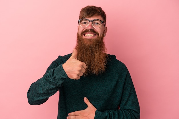 Un jeune homme de race blanche au gingembre avec une longue barbe isolée sur fond rose touche le ventre, sourit doucement, mange et satisfait le concept.