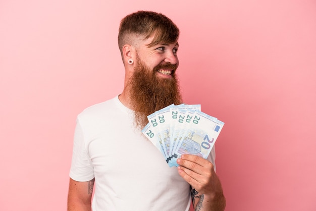 Jeune homme de race blanche au gingembre avec une longue barbe tenant des billets de banque isolés sur fond rose regarde de côté souriant, joyeux et agréable.