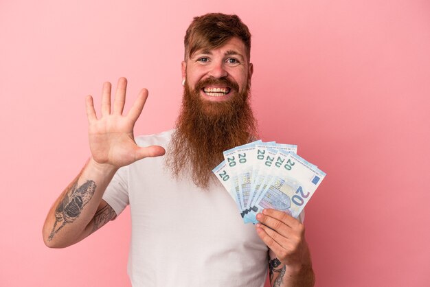 Jeune homme de race blanche au gingembre avec une longue barbe tenant des billets de banque isolés sur fond rose souriant joyeux montrant le numéro cinq avec les doigts.