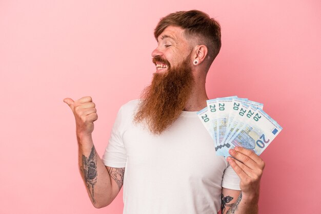 Jeune homme de race blanche au gingembre avec une longue barbe tenant des billets de banque isolés sur des points de fond rose avec le pouce loin, riant et insouciant.