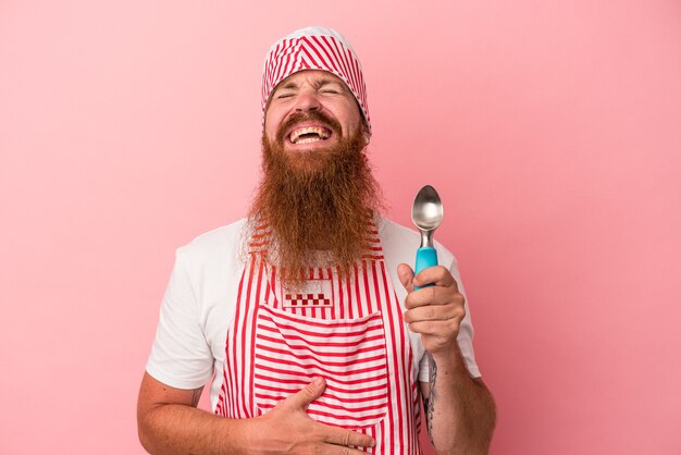 Jeune homme de race blanche au gingembre avec une longue barbe tenant une boule isolée sur fond rose rit fort en gardant la main sur la poitrine.