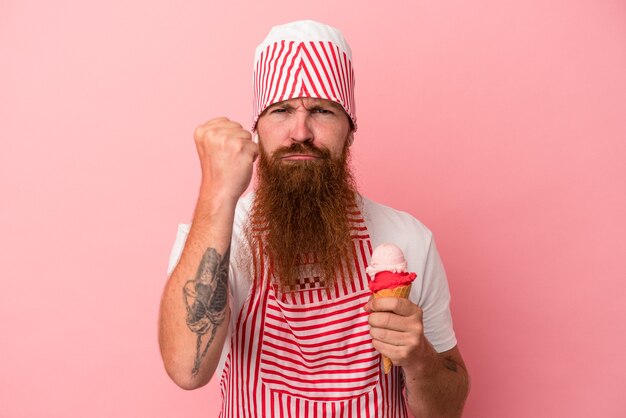 Photo jeune homme de race blanche au gingembre avec une longue barbe tenant une glace isolée sur fond rose montrant le poing à la caméra, expression faciale agressive.