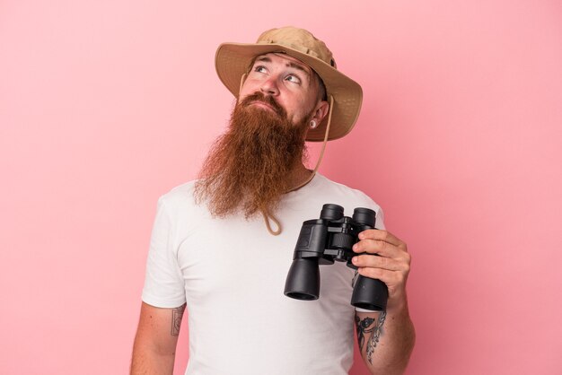 Jeune homme de race blanche au gingembre avec une longue barbe tenant des jumelles isolées sur fond rose rêvant d'atteindre des objectifs et des objectifs