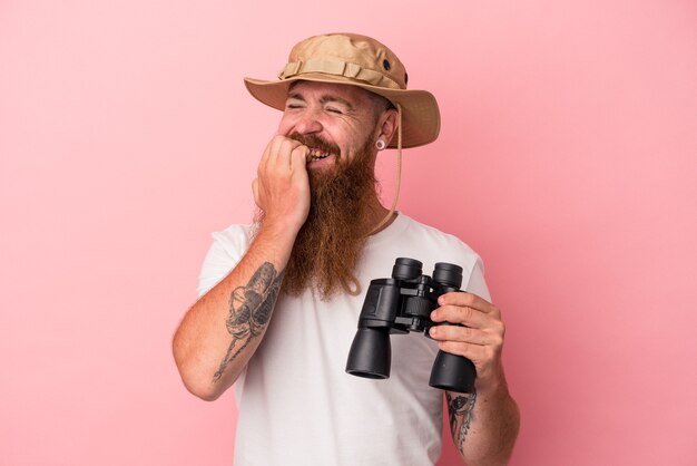 Jeune homme de race blanche au gingembre avec une longue barbe tenant des jumelles isolées sur fond rose se rongeant les ongles, nerveux et très anxieux.