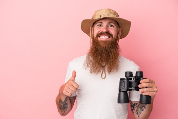 Jeune homme de race blanche au gingembre avec une longue barbe tenant des jumelles isolées sur fond rose souriant et levant le pouce vers le haut