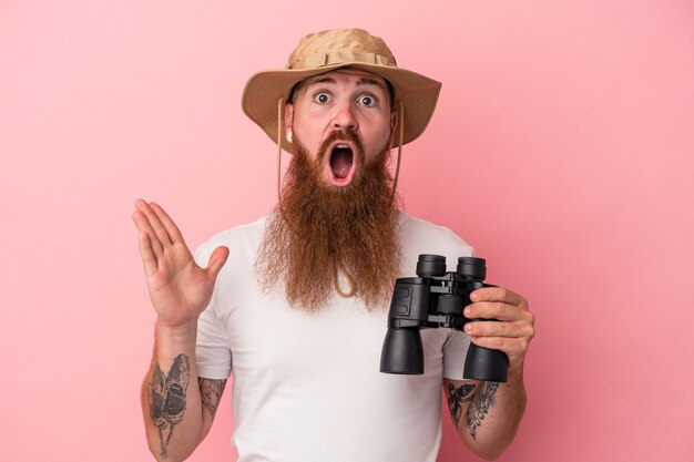 Jeune homme de race blanche au gingembre avec une longue barbe tenant des jumelles isolées sur fond rose surpris et choqué.