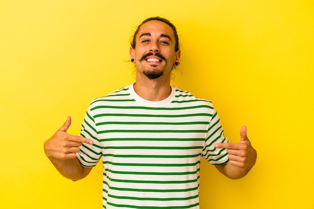 Jeune homme de race blanche aux cheveux longs isolé sur fond jaune pointe vers le bas avec les doigts, sentiment positif.