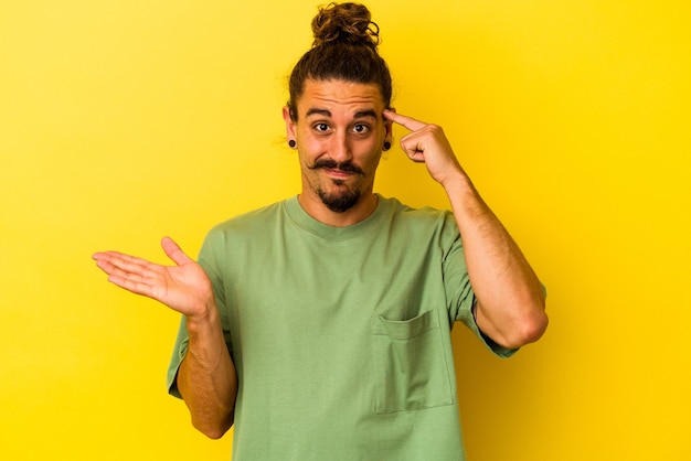 Jeune homme de race blanche aux cheveux longs isolé sur fond jaune tenant et montrant un produit à portée de main.