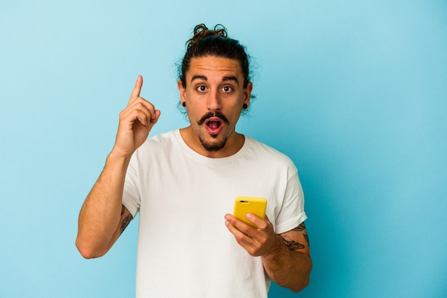 Jeune homme de race blanche aux cheveux longs tenant un téléphone portable isolé sur fond bleu ayant une idée, un concept d'inspiration.