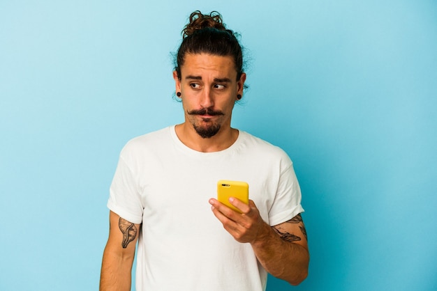 Jeune homme de race blanche aux cheveux longs tenant un téléphone portable isolé sur fond bleu confus, se sent dubitatif et incertain.