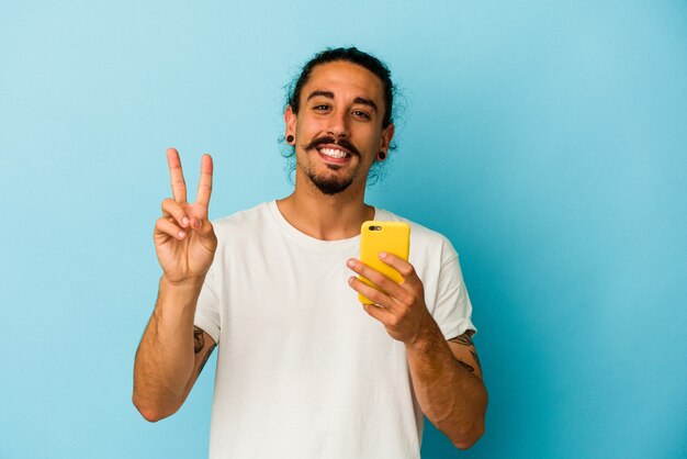 Jeune homme de race blanche aux cheveux longs tenant un téléphone portable isolé sur fond bleu joyeux et insouciant montrant un symbole de paix avec les doigts.