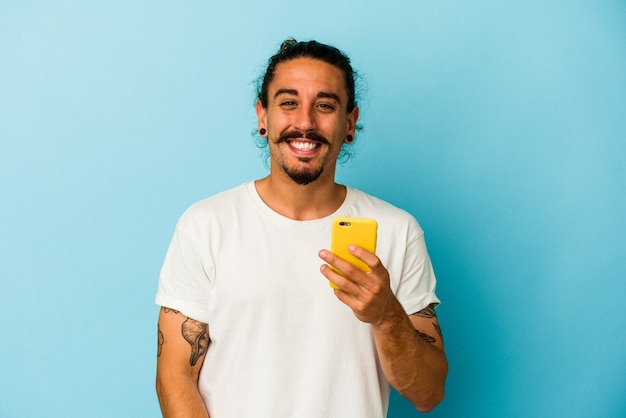 Jeune homme de race blanche aux cheveux longs tenant un téléphone portable isolé sur fond bleu en riant et en s'amusant.