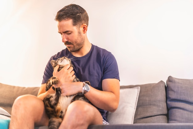 Un jeune homme de race blanche aux cheveux noirs dans un T-shirt bleu et un short jouant à la maison avec son beau chat domestique gris et blanc. Le meilleur ami de l'homme chat. Caressant le joli chat assis sur le canapé