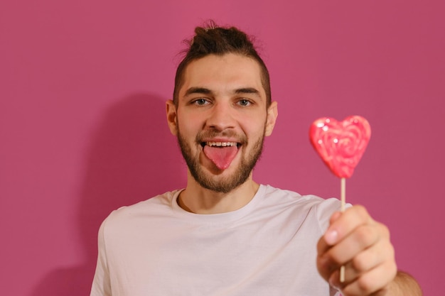 Jeune homme de race blanche avec barbe montre la langue et taquine la sucette rouge douce en forme de coeur