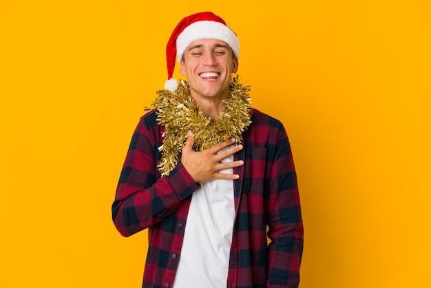 Jeune homme de race blanche avec un chapeau de Noël tenant un cadeau isolé sur un mur jaune, éclate de rire en gardant la main sur la poitrine.